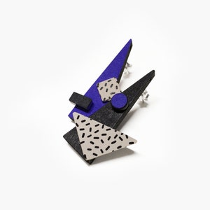May include: Geometric stud earrings featuring a layered design with black, blue, and gray triangles and shapes. The earrings have a speckled pattern on the gray triangle and square, and a small blue circle. The earrings have silver posts.