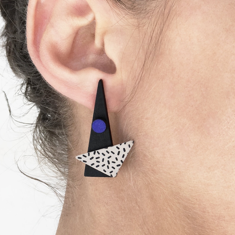 May include: A black and white geometric earring with a triangular shape. The earring features a black triangle with a small blue circle and a white triangle with black speckles. The earring is worn on the ear.