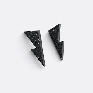 Puede incluir: Un par de pendientes negros con forma de rayo y motas brillantes. Los pendientes son de un material oscuro y texturizado, sobre un fondo blanco. Diseño geométrico único.
