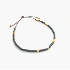 以下が含まれることがあります： スライドノットクロージャーが付いたグレーとゴールドのビーズブレスレット。ブレスレットは、小さな円筒形のビーズと、その間に散りばめられたゴールドの正方形のビーズでできています。