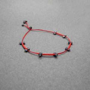 Op de afbeelding: Een rode armband met kleine, donkergrijze kralen gelijkmatig verdeeld over de draad. De armband heeft een verstelbare schuifknoopsluiting. De kralen zijn donkergrijs van kleur en lijken van glad materiaal te zijn.