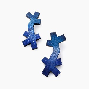 May include: A pair of earrings featuring an irregular, multi-armed cross shape. The surface displays a gradient of deep blue to lighter blue, speckled with tiny white dots that create a starry night effect across the textured material.