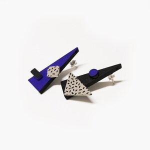 May include: A pair of geometric stud earrings. One earring is a blue triangle with a black rectangle and a square with a black dash pattern. The other earring is a black triangle with a gray triangle with a black dash pattern and a blue circle.