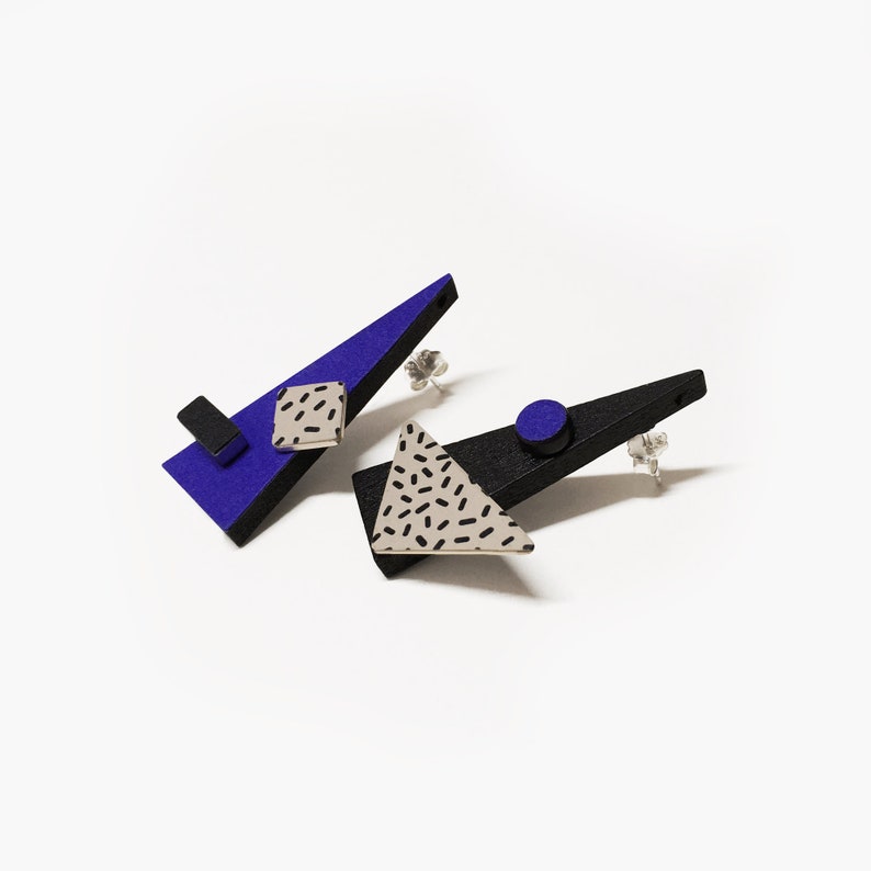 May include: A pair of geometric stud earrings. One earring is a blue triangle with a black rectangle and a square with a black dash pattern. The other earring is a black triangle with a gray triangle with a black dash pattern and a blue circle.