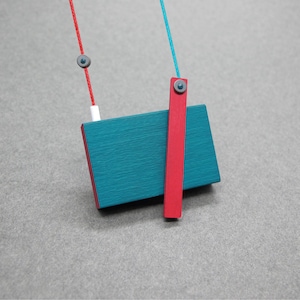 May include: A modern necklace featuring a teal rectangular pendant with a red accent and a vertical red bar. The pendant is suspended from a red and blue cord with small gray beads. The design is minimalist and geometric.
