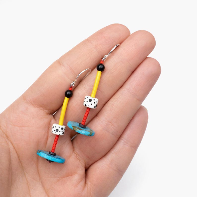 K&ouml;nnte beinhalten: Ein Paar handgefertigte Ohrringe mit einem lebendigen Design. Jeder Ohrring hat eine t&uuml;rkisfarbene Scheibe, eine wei&szlig;e Perle mit schwarzen Punkten, einen gelben Stab und einen roten Stab. Die Ohrringe werden in einer Hand gehalten.