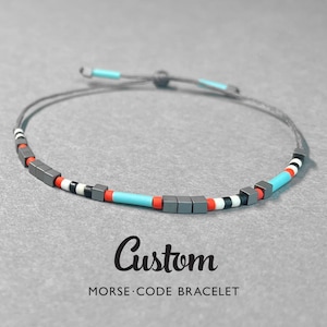 Può includere: Un braccialetto con codice Morse personalizzato con cordino grigio e chiusura regolabile. Il braccialetto presenta perline grigie, rosse, bianche, nere e turchesi. Il testo "Custom MORSE CODE BRACELET" è visualizzato sotto.