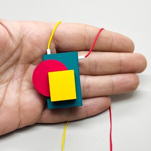 Hand holding a colorful geometric Bauhaus style pendant necklace, abstract wooden statement jewelry with bold Memphis inspired shapes, modern avant garde necklace photographed in natural soft light, artistic contemporary fashion accessory close-up in hand