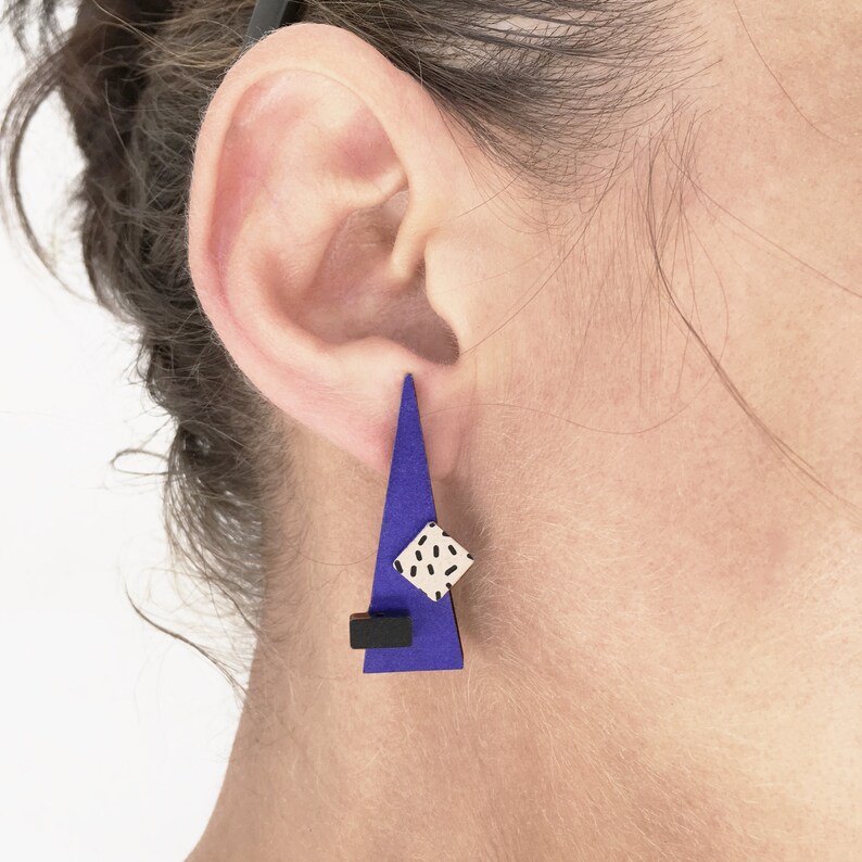May include: A geometric earring with a blue triangle shape, a small black rectangle, and a white square with black speckles. The earring is worn on the ear.