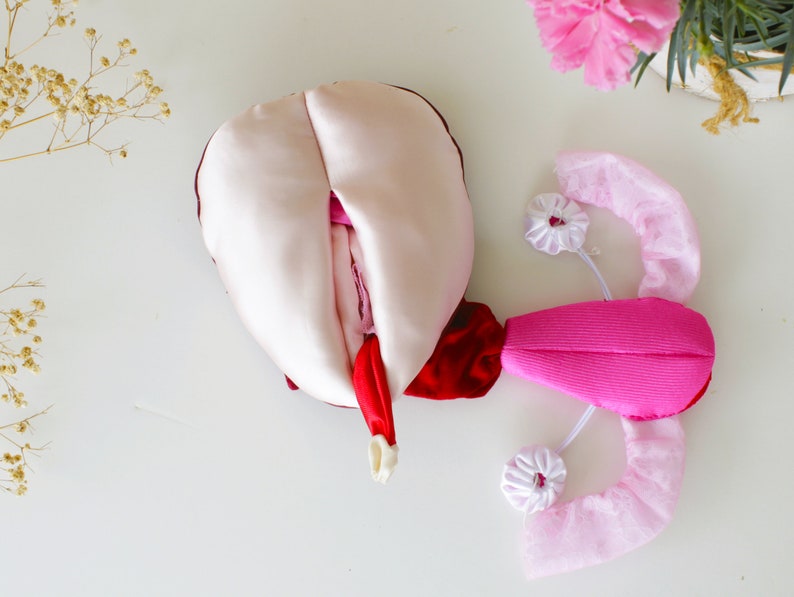 May include: A pink and red fabric flower with a white satin center. The flower is open and has a ruffled edge. The flower is surrounded by a white tulle fabric.