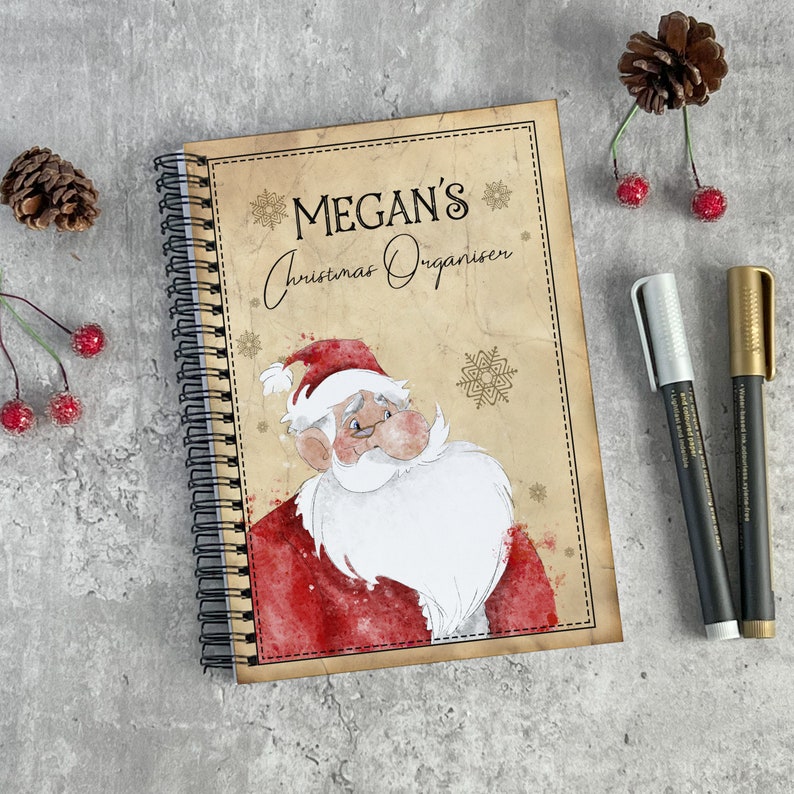 May include: A brown spiral bound notebook with a vintage-style cover featuring a cartoon Santa Claus with a red and white suit and a white beard. The cover also has the text "MEGAN'S Christmas Organiser" in gold lettering.