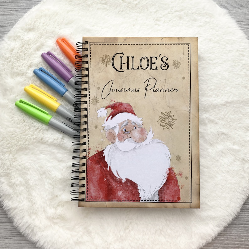 May include: A spiral bound Christmas planner with a vintage-style cover featuring a cartoon Santa Claus with a red hat and white beard. The cover also has the text "CHLOE'S Christmas Planner" and snowflakes.