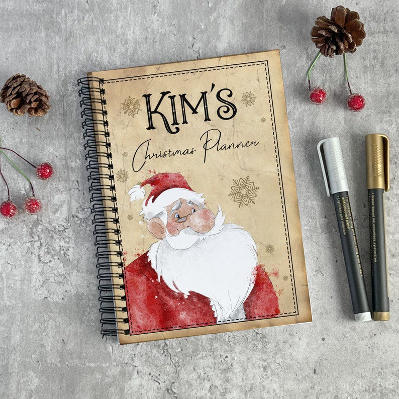 May include: A brown spiral bound notebook with a Christmas theme. The cover features a cartoon Santa Claus with a white beard and red hat. The text "KIM'S Christmas Planner" is printed in gold on the cover.