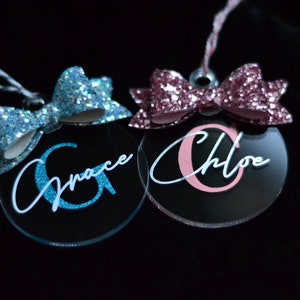 Peut inclure: Deux décorations de Noël personnalisées avec des nœuds pailletés. Un ornement est bleu avec le nom "Grace" et l'autre est rose avec le nom "Chloe".