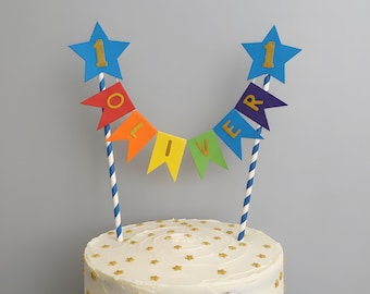 May include: A cake topper featuring two blue star cutouts with the number 1 in gold, connected by a string of colorful pennant flags spelling the name OLIVER in gold letters, supported by two blue and white striped paper straws on a white cake.