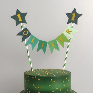 May include: A green cake decorated with gold star sprinkles and a banner reading "OLIVER". The cake topper features two dark green star decorations with the number "1" in gold. The banner flags are in shades of green and gold.