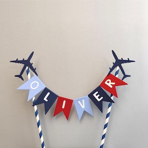 May include: A cake topper with the name "OLIVER" displayed on a banner of blue and red pennants. Two dark blue airplane silhouettes are positioned on either side of the banner. The topper is supported by two blue and white striped straws.