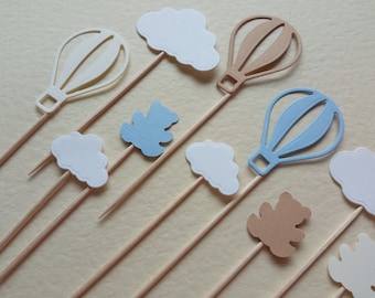 May include: A set of nine paper cupcake toppers featuring hot air balloons, clouds, and teddy bears. The toppers are in shades of blue, brown, and white and are attached to wooden skewers.