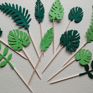 May include: Assortment of green paper tropical leaf cake toppers on wooden skewers. The leaves are in various shapes and sizes, including monstera, fern, and palm leaves.