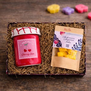 Op de afbeelding: Een geschenkdoos met een rode keramische pot met een wit deksel met rood ruitpatroon en een etiket met de tekst Sugar Sweet Cherry Jam, naast een bruin papieren zakje met het label Bohemian Summer Tales gevuld met gele waxmelts.