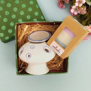 以下が含まれることがあります： 青いアクセントが付いた白いセラミック製のキノコ型オイルウォーマー。緑色の水玉模様のギフトボックスに入っており、小さなキャンドルの箱も付いています。箱には藁のような詰め物が含まれており、ピンクの花の隣にあります。