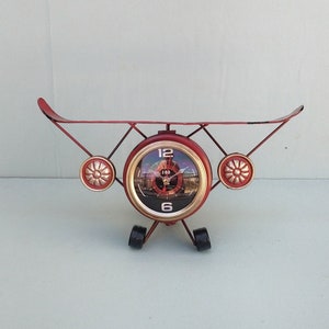 May include: Red metal aeroplane clock with gold accents. The clock face has black Roman numeral markings and a black and white image of an aeroplane in the centre.
