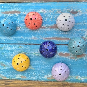 Pode incluir: Sete esferas decorativas de cerâmica em várias cores, incluindo azul, laranja, branco, amarelo, roxo e verde-azulado. Cada esfera tem uma superfície texturizada com orifícios espaçados, semelhantes a ouriços-do-mar. As esferas estão dispostas sobre uma superfície de madeira azul desgastada.