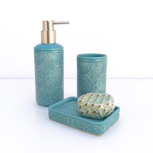 Puede incluir: Un juego de baño turquesa con un patrón floral. El juego incluye un dispensador de jabón con bomba dorada, un vaso, una jabonera y una pastilla de jabón con diseño geométrico. Los artículos están dispuestos sobre una superficie blanca.