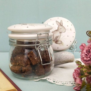 Op de afbeelding: Een glazen pot met een keramische deksel, gevuld met bruine koekjes. De deksel en een aparte keramische schijf hebben een konijnenillustratie. Een metalen sluiting zet de pot vast. Roze bloemen en een boek staan op de achtergrond.