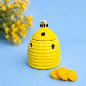Puede incluir: Un tarro de cerámica amarillo brillante con forma de colmena y tapa extraíble que presenta una pequeña figura de abeja negra y amarilla en la parte superior. El tarro tiene aberturas hexagonales y tres pequeñas ceras redondas.