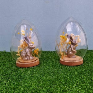 Puede incluir: Dos cúpulas de cristal en forma de huevo, cada una con una pequeña figura de conejo de metal plateado y flores amarillas secas. Las cúpulas están montadas sobre bases circulares de madera, ofreciendo una vista clara del contenido.
