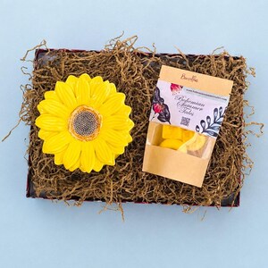 Peut inclure: Une boîte cadeau remplie de papier déchiqueté marron contenant un plat en céramique jaune vif en forme de tournesol et un petit sachet en papier kraft étiqueté Banoffee et Bohemian Summer Tales avec une fenêtre montrant des cires parfumées.