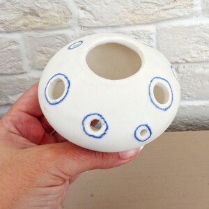 May include: A white ceramic flower frog with a rounded, dome-like shape. The surface features several circular openings, each outlined with a hand-painted blue ring. The central opening is larger than the surrounding smaller holes.