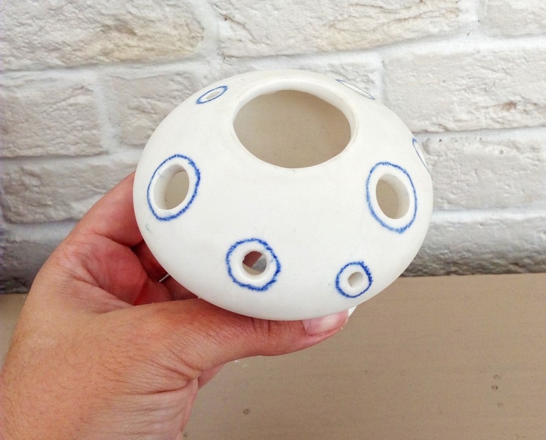 May include: A white ceramic flower frog with a rounded, dome-like shape. The surface features several circular openings, each outlined with a hand-painted blue ring. The central opening is larger than the surrounding smaller holes.