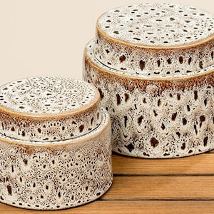 Puede incluir: Dos tarros de cerámica con tapa, con un patrón moteado en crema y marrón. Los tarros son cilíndricos con una tapa redondeada. Las tapas y los bordes tienen un esmalte marrón. Los tarros están apilados sobre una superficie de madera.