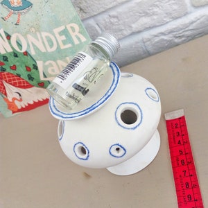 May include: A white ceramic flower frog with blue-rimmed circular openings, topped with a small clear glass bottle featuring a barcode and text. A red measuring tape is positioned alongside, showing a length of approximately 10.5 centimeters.