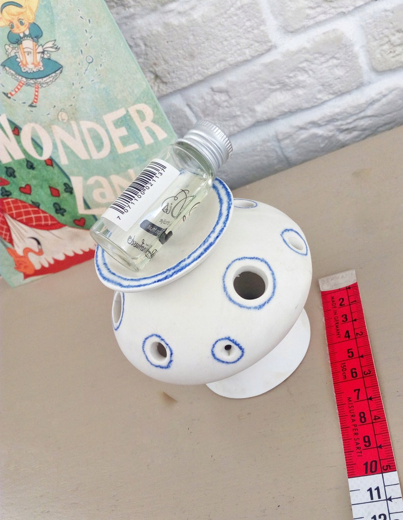 May include: A white ceramic flower frog with blue-rimmed circular openings, topped with a small clear glass bottle featuring a barcode and text. A red measuring tape is positioned alongside, showing a length of approximately 10.5 centimeters.