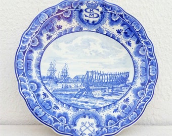 May include: A round ceramic plate with a scalloped edge featuring a blue and white transferware scene of a shipyard with sailing ships and a wooden hull under construction. The top displays a crown above the letters KM, and the bottom shows anchors.