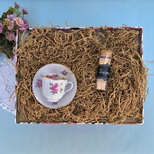 Könnte beinhalten: Eine Geschenkbox mit einer weißen Tasse und Untertasse mit rosa Rosen und goldenen Verzierungen. Ein kleines Glasfläschchen mit Streichhölzern, beschriftet mit dem Wort "Σπίρτα", ist ebenfalls enthalten. Die Box ist mit braunem Papier gefüllt und hat ein Blumen- und Streifenmuster.
