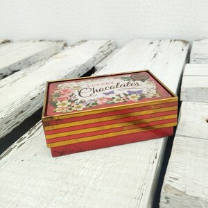 May include: A red and gold tin box with a floral design and the text "Luxury Chocolates" on the lid. The box has a striped pattern on the sides.