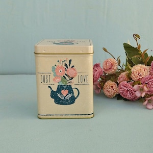 Può includere: Una scatola di metallo rettangolare color crema con coperchio. La parte anteriore presenta un'illustrazione di una teiera blu scuro riempita con fiori rosa e arancioni, un piccolo cuore e il testo "JUST LOVE" stampato in nero ai lati della teiera.