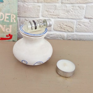 May include: A white ceramic oil burner with a blue-rimmed lid and blue circular accents around the base. A small glass bottle labeled "Dis Duftolie Passion fruit Kammerhuset" with a silver cap rests on top, and a white tea light candle sits nearby.