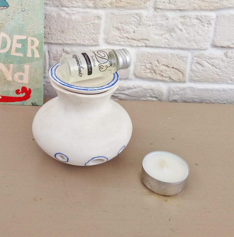 May include: A white ceramic oil burner with a blue-rimmed lid and blue circular accents around the base. A small glass bottle labeled "Dis Duftolie Passion fruit Kammerhuset" with a silver cap rests on top, and a white tea light candle sits nearby.
