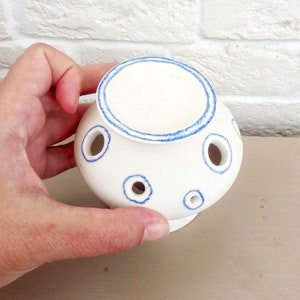 May include: A white ceramic flower frog with a rounded body and a flat top. The piece features multiple circular openings for stems, each outlined with a hand-painted blue ring. A matching blue ring circles the top edge of the ceramic vessel.