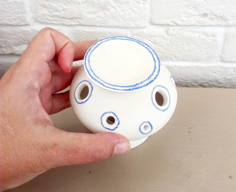 May include: A white ceramic flower frog with a rounded body and a flat top. The piece features multiple circular openings for stems, each outlined with a hand-painted blue ring. A matching blue ring circles the top edge of the ceramic vessel.