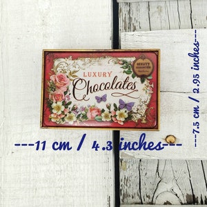 May include: A red and gold metal tin with a floral design and the words "LUXURY Chocolates" printed on it. The tin is 4.3 inches wide and 2.95 inches tall.