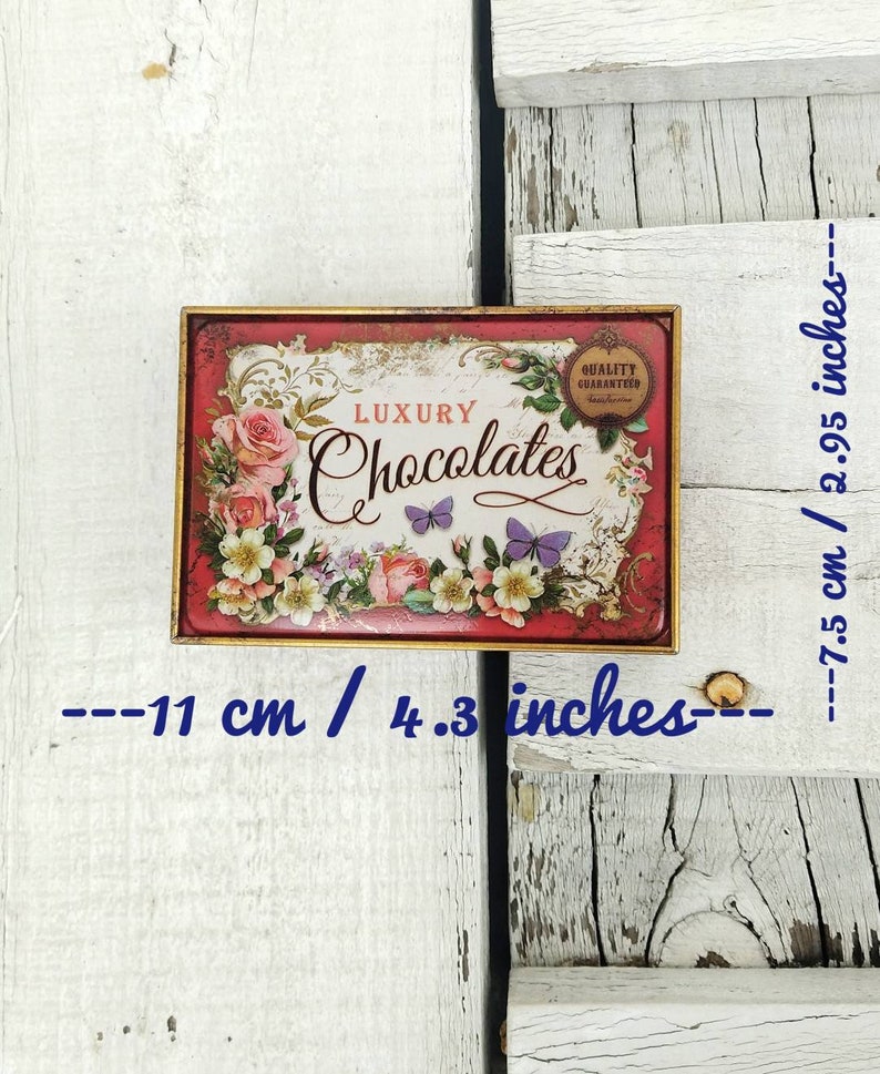 May include: A red and gold metal tin with a floral design and the words "LUXURY Chocolates" printed on it. The tin is 4.3 inches wide and 2.95 inches tall.