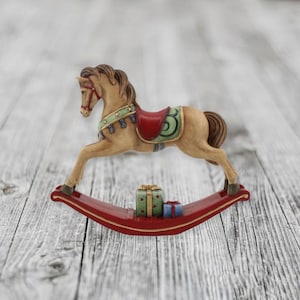 May include: A tan rocking horse figurine with a brown mane and tail, featuring a red saddle, a green blanket with gold stars, and a red bridle. The curved base is red and holds two small gift boxes, one green and one blue, between the rockers.
