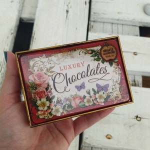 May include: A red and gold tin box with a floral design and the text "Luxury Chocolates" and "Quality Guaranteed".