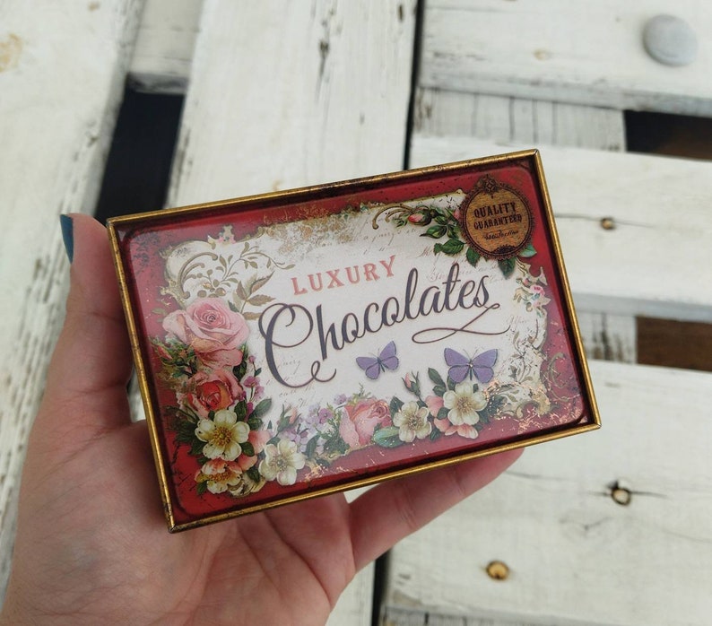 May include: A red and gold tin box with a floral design and the text "Luxury Chocolates" and "Quality Guaranteed".